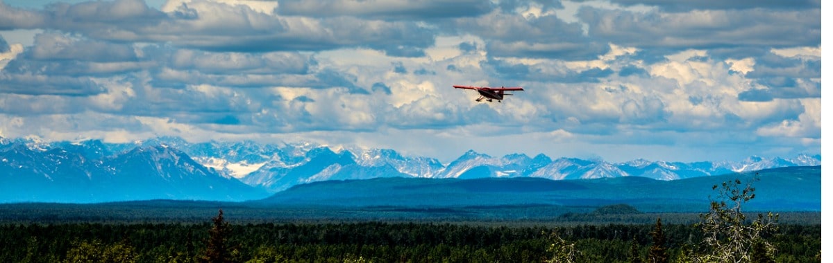 Small red plane flying over Alaska, with snow capped mountains in the background